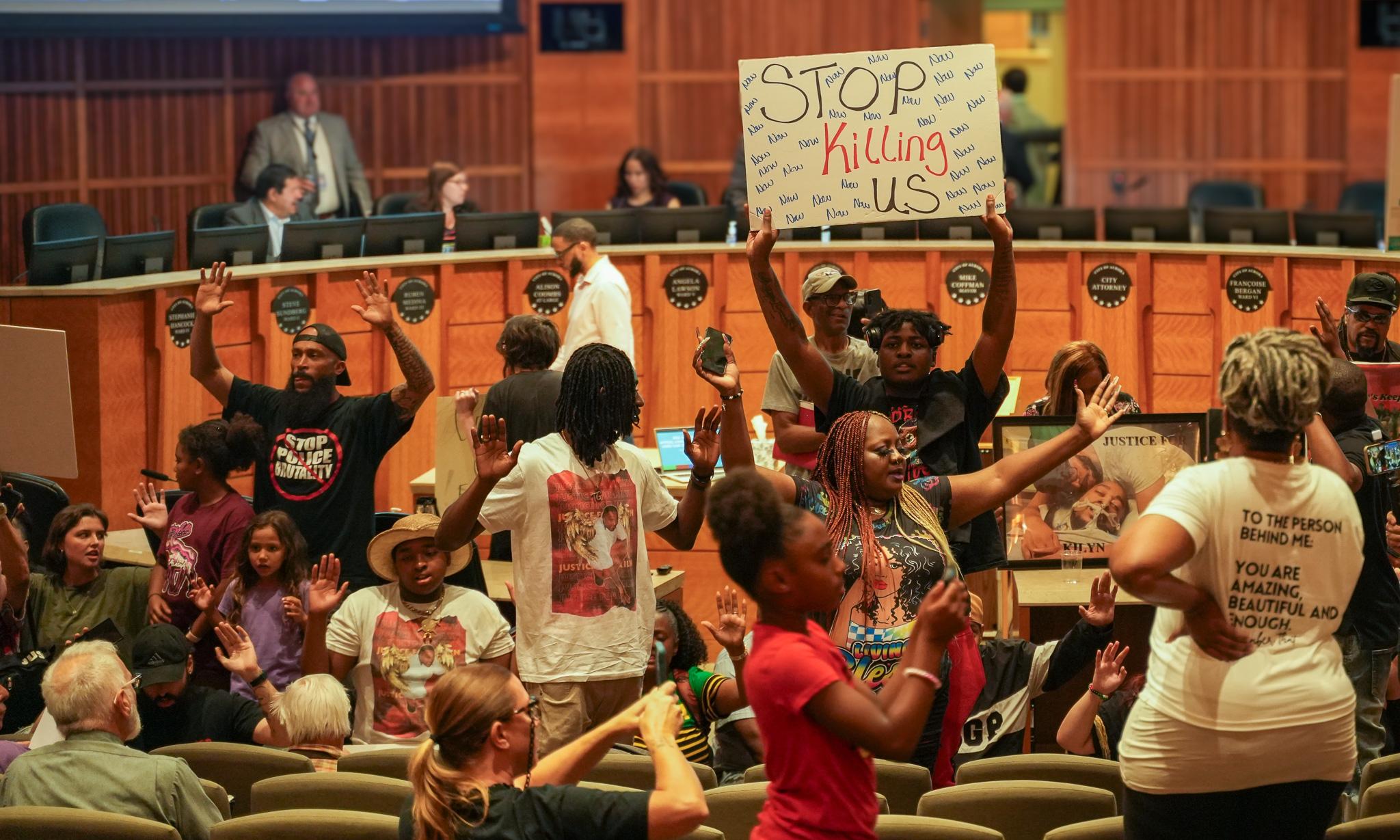 Security officer watches from doorway as community members sit on the floor of council chambers during protest