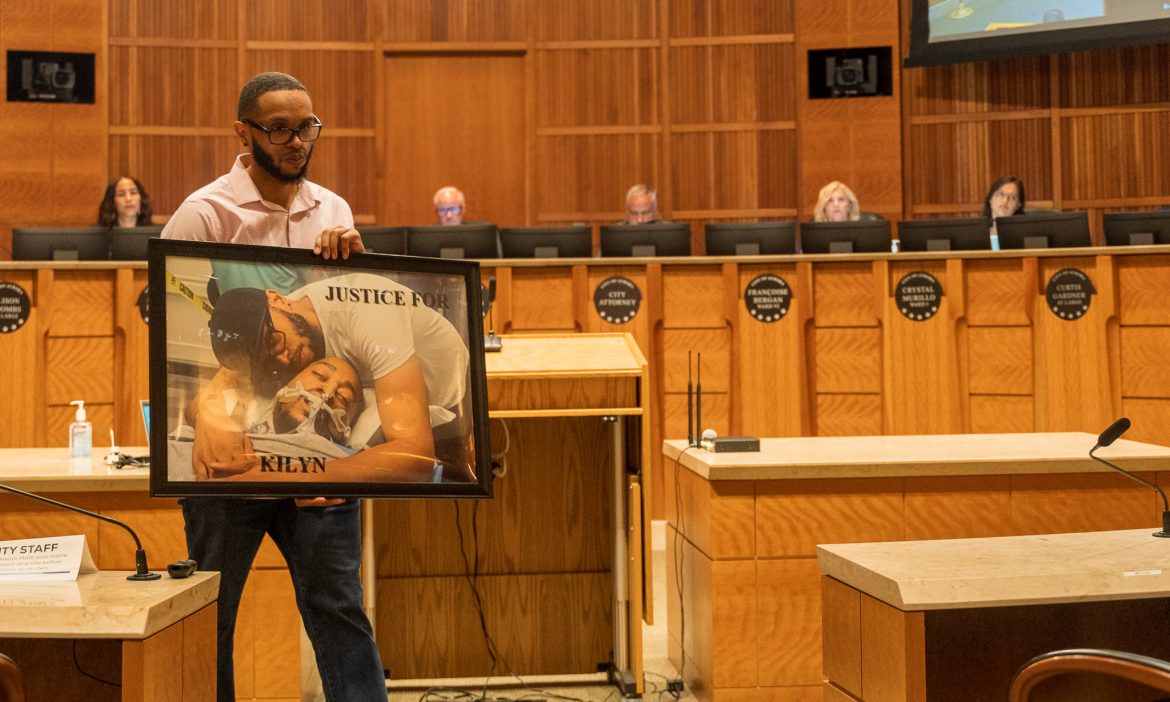 A man holds a large framed photo reading 'Justice for Kilyn' before the Aurora City Council dais