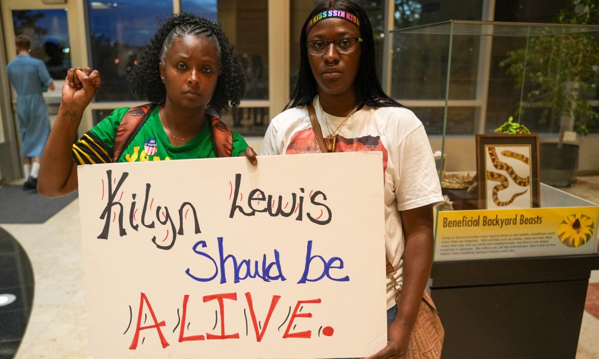 Two women holding a sign reading 'Kilyn Lewis Should Be ALIVE' outside Aurora City Council
